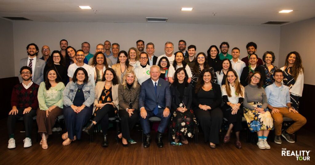 Grupo de pessoas em foto coletiva em ambiente interno. Ao centro, sentado na primeira fileira, está Al Gore, ex-vice-presidente dos Estados Unidos e Prêmio Nobel da Paz. As demais pessoas, em pé ou sentadas ao redor, posam sorridentes para a câmera. No canto inferior direito, aparece o logotipo escrito 'The Reality Tour'.